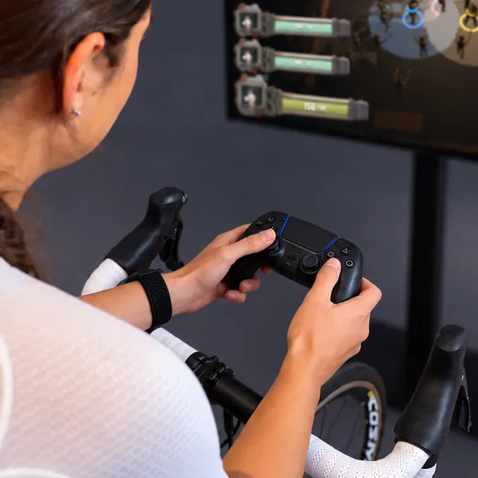 A rider on a smart trainer holding a PlayStation controller, a RYDR game on the screen ahead