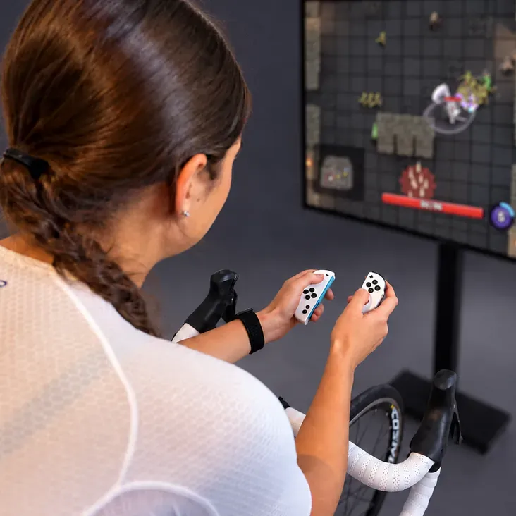 A rider on a smart trainer holding a Switch Joy-Con in each hand, playing a game on a big screen